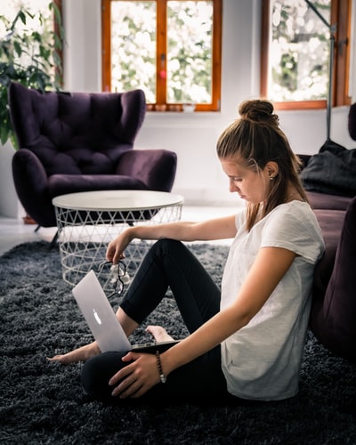 Dressed in a white T-shirt and black leggings lady sitting area rug, facing the silver MacBook
